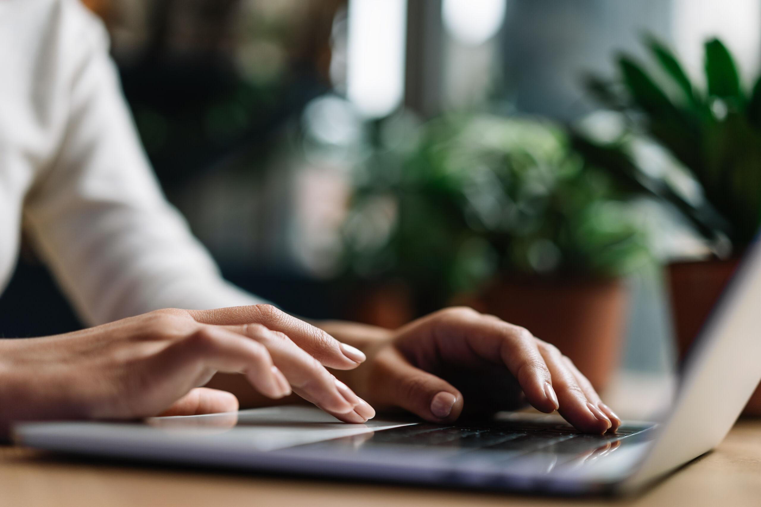 Close-up of a person typing on a laptop keyboard, working at a desk with green plants blurred in the background — representing focus, productivity, or completing an online task.