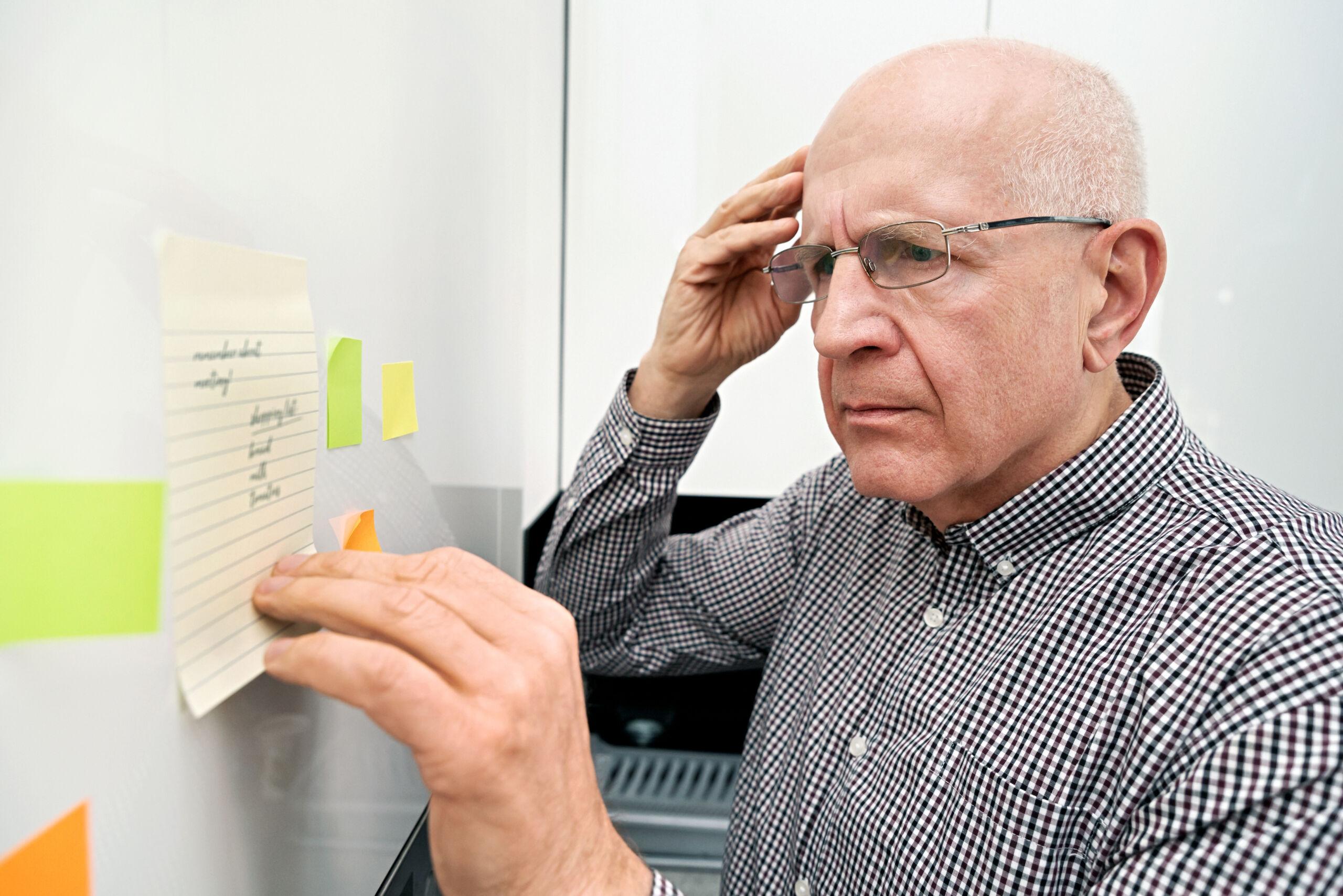 An older man wearing glasses and a checkered shirt looks intently at a handwritten note taped to a wall or refrigerator, appearing focused and slightly puzzled as he reads reminders surrounded by colorful sticky notes — illustrating memory difficulties or forgetfulness.