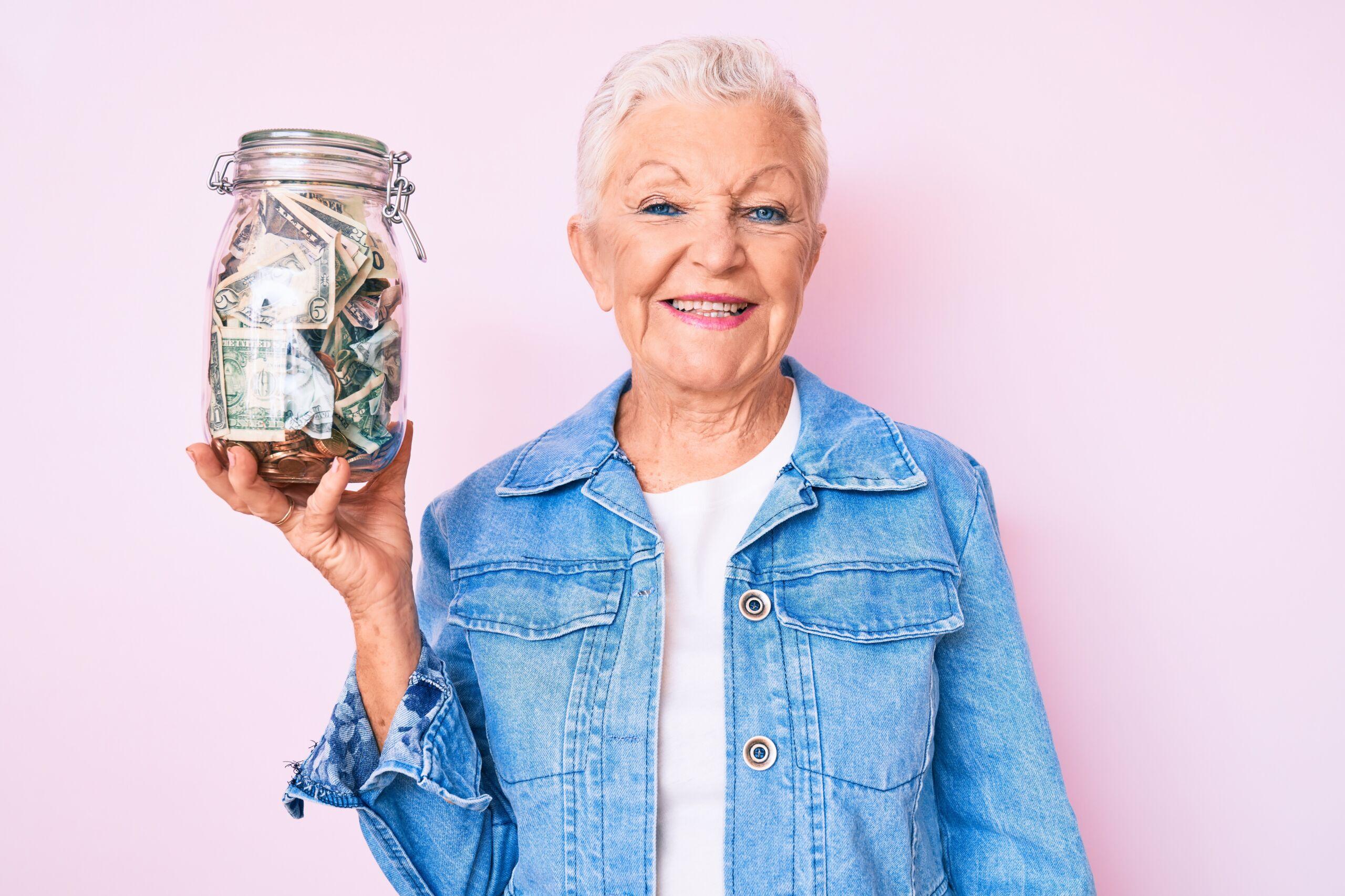 Smiling older woman holding a large jar filled with cash against a pink background.