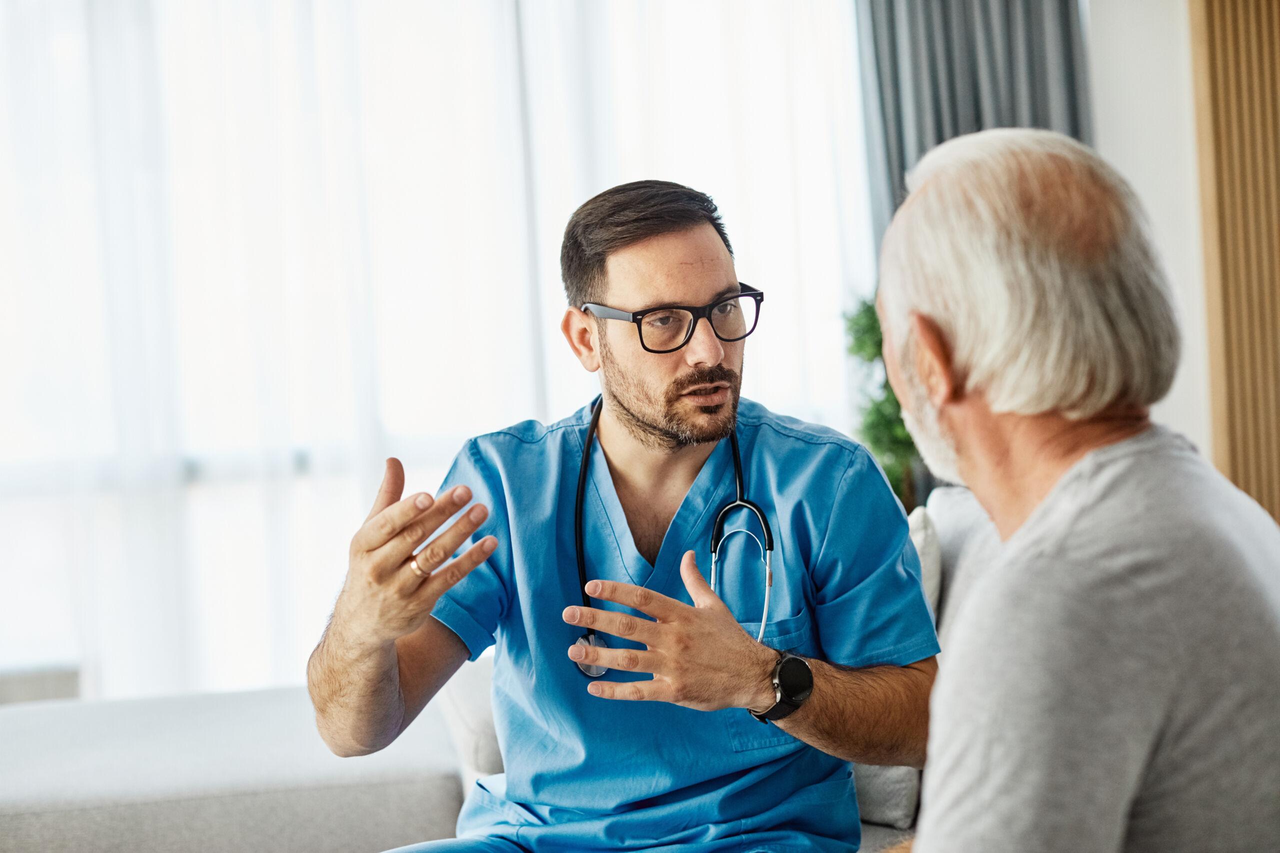 A healthcare professional wearing blue scrubs and a stethoscope speaks with an older male patient in a bright medical setting, explaining something with hand gestures — representing a doctor–patient consultation or discussion about health concerns.