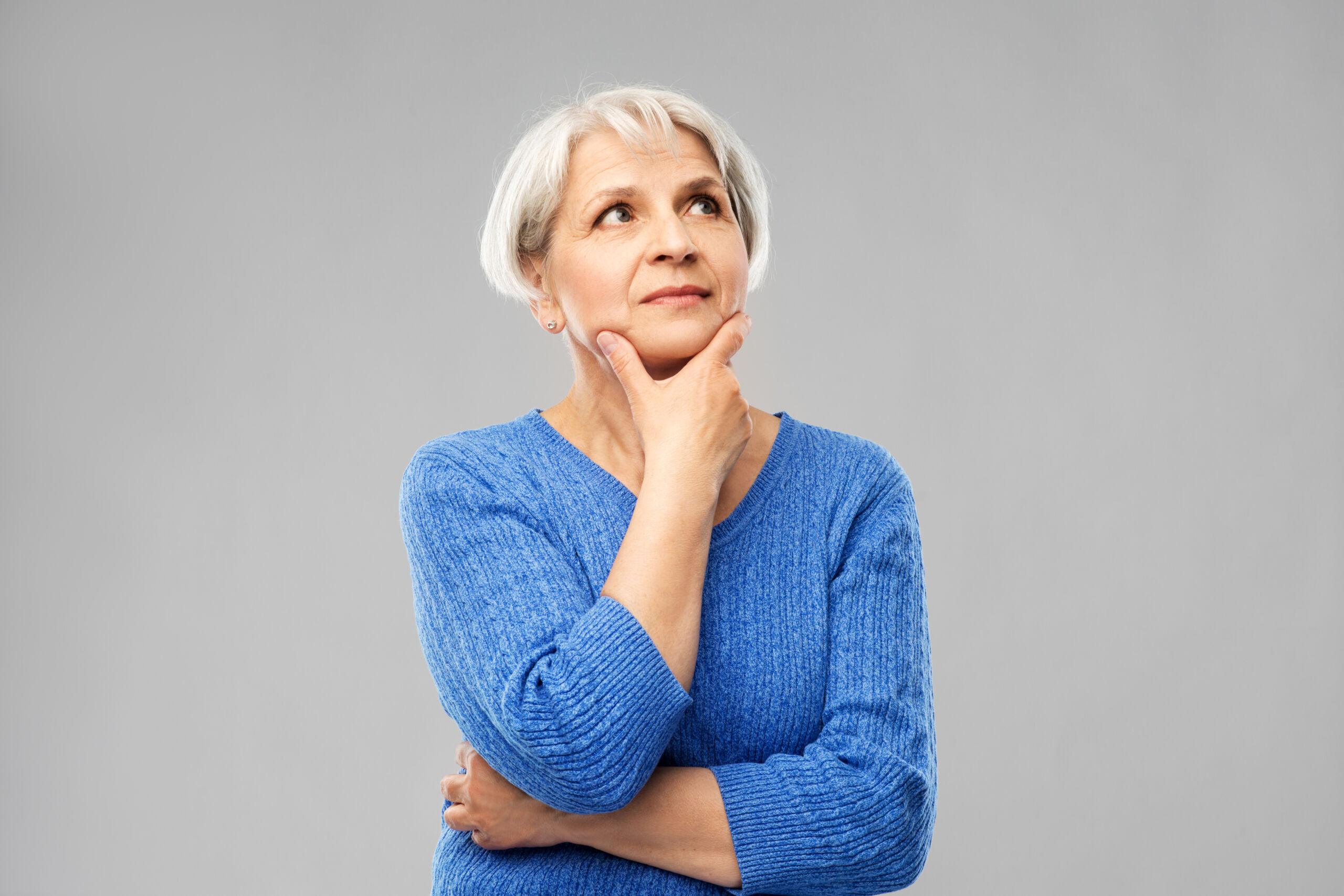 Older woman with short gray hair wearing a blue sweater, looking up thoughtfully with her hand on her chin against a plain light gray background