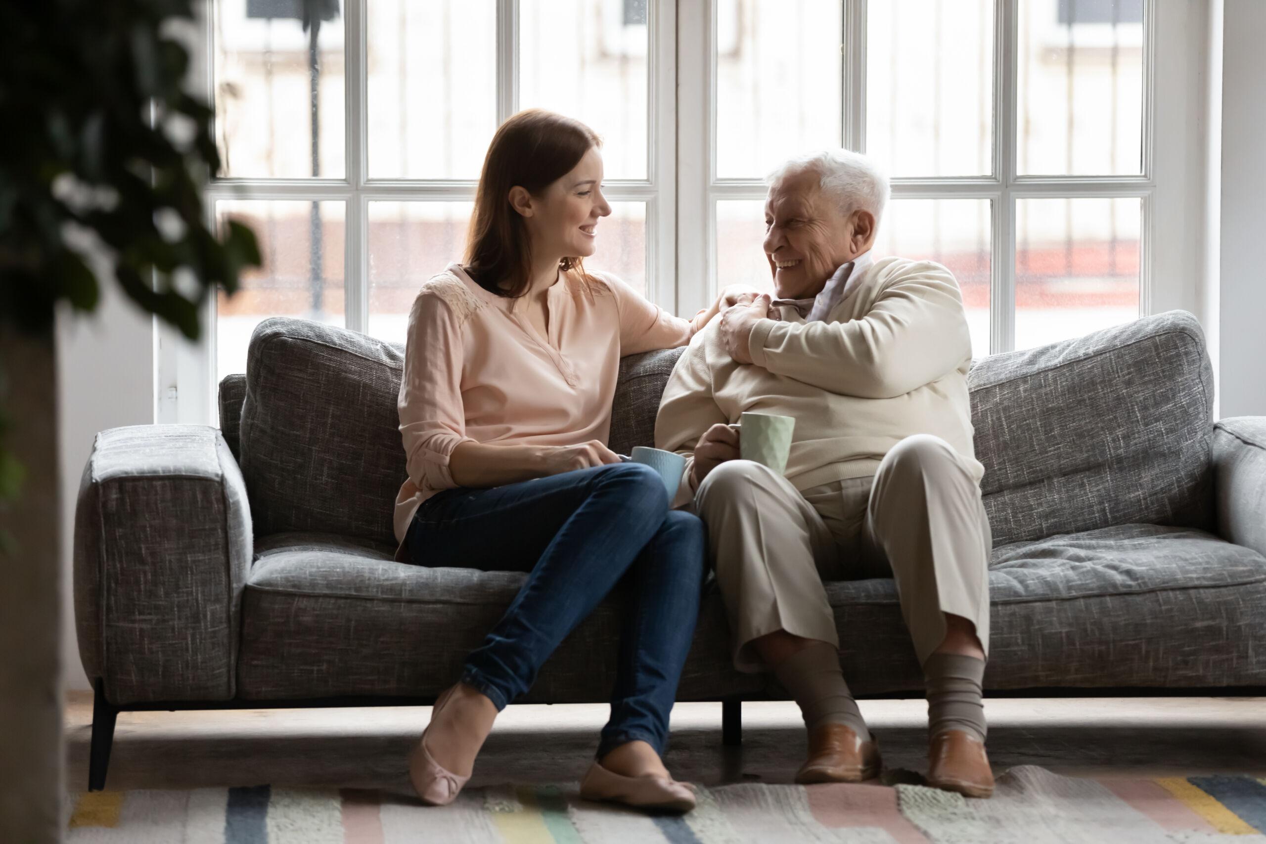 Adult daughter sitting on a couch talking warmly with her elderly father in a bright living room, sharing a supportive conversation.